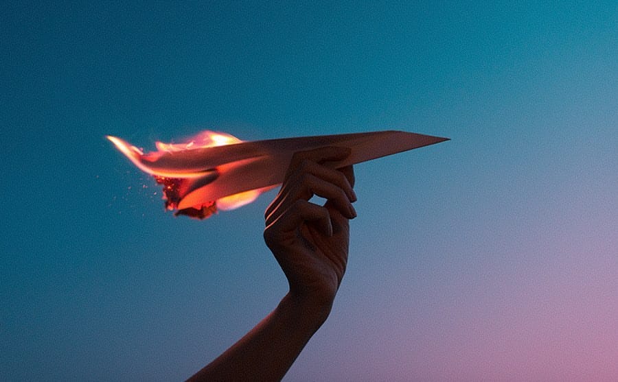 Main qui s'apprête à lancer un avion en papier qui prend feu, ciel de soleil couchant en fond