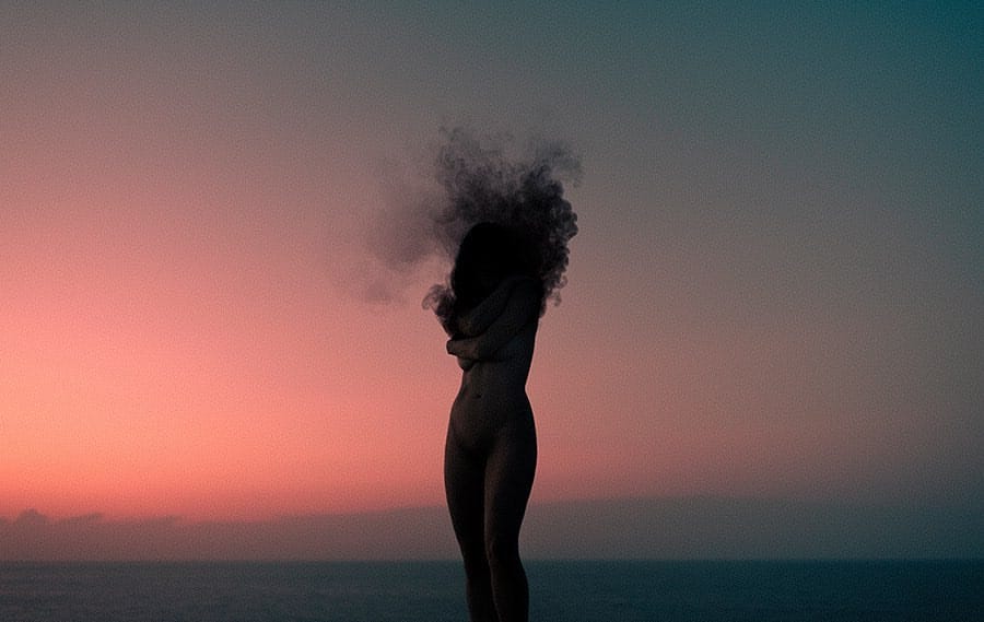 Une jeune femme est debout, nue, au soleil couchant devant la mer. à la place de sa tête il y a une dense fumée sombre