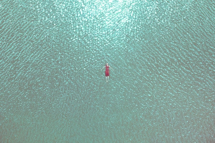 Photo prise de haut, on voit une femme habillée en rouge allongée sur le sable recouvert par un fond d'eau