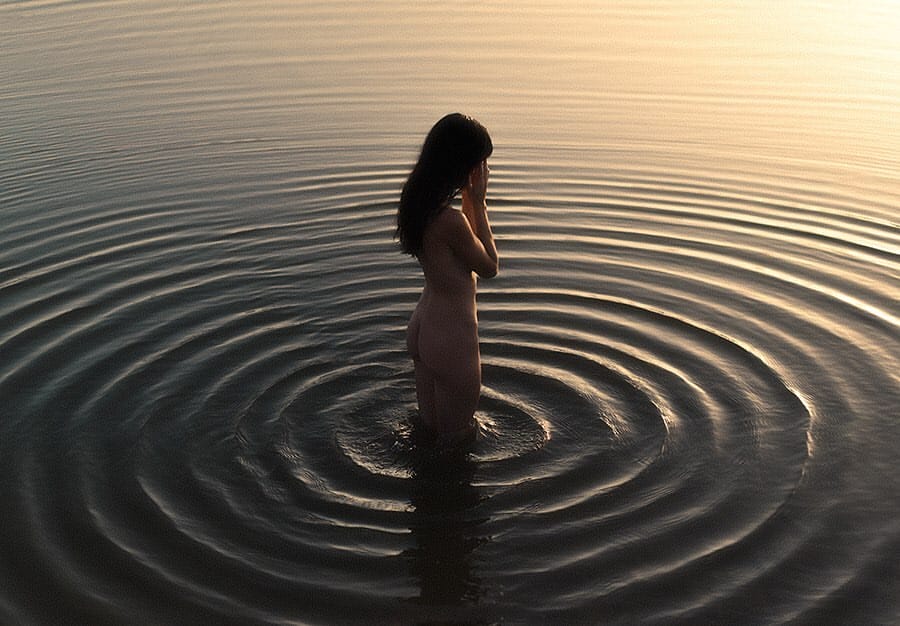 Une jeune femme nue est debout, faisant presque de dos à nous, les jambes dans l'eau. L'eau décrit des cercles autour d'elle