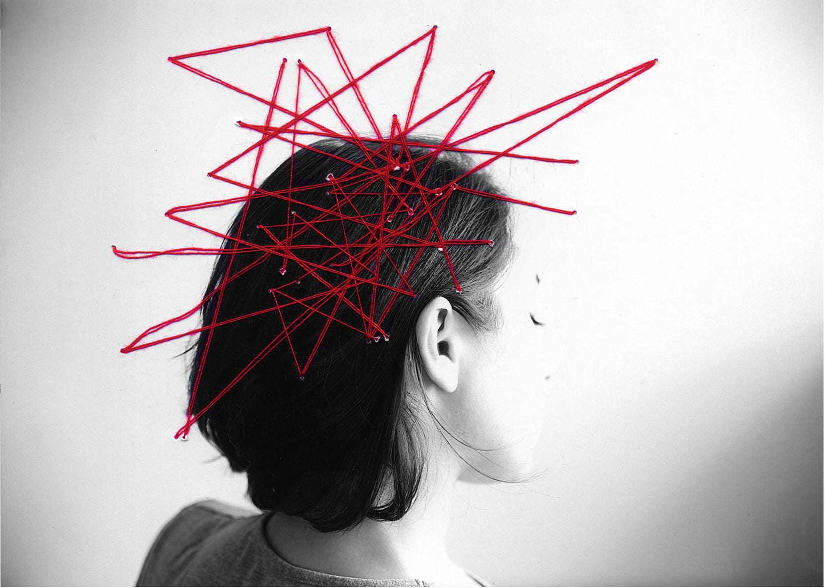 Photographie en noir et blanc d'une femme de dos. Des fils rouges sont épinglés sur la photo, sur la tête de la femme