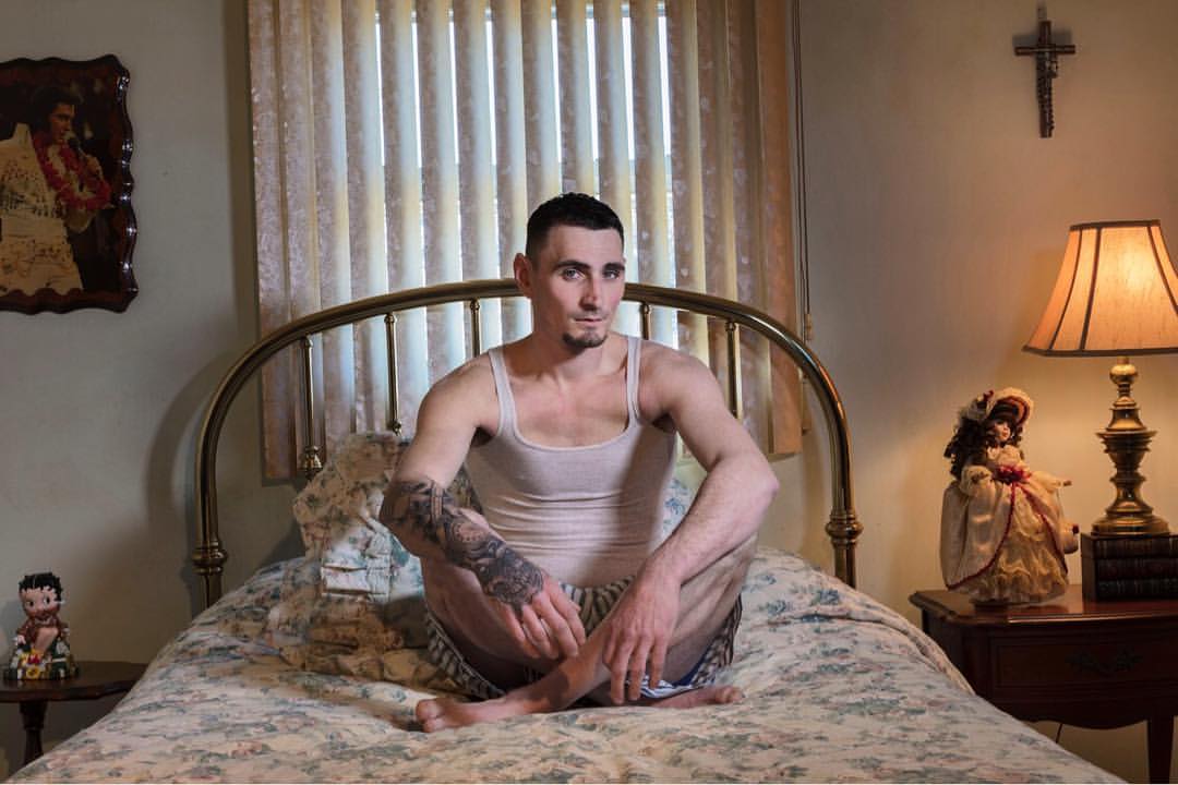 Homme assis sur son lit en tailleur, en sous vêtements. Son bras est tatoué. Danna Singer