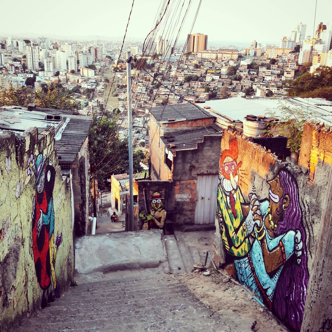 Vue des favelas brésiliennes. Sur un mur, deux personnages bariolés dansent une salsa, sur un autre, on distingue un personnage mi-femme âgée, mo-coccinelle. En fond, tag d'un un criquet déguisé en chasseur.