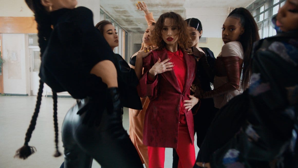 Groupe de femmes, entourent le personnage principal vêtue de rouge comme pour la protéger. Elles dansent ensemble.