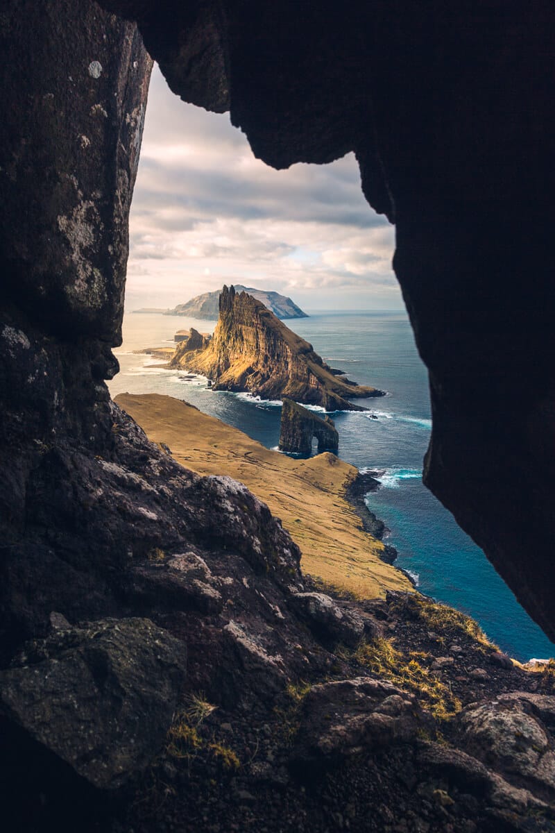 Point de vue entre deux rochers d'une plaine en bord de mer avec massifs rocheux et d'un pic sortant de l'eau turquoise. Nuages blancs dans le ciel.