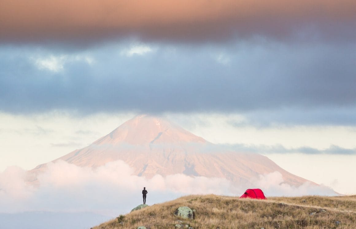 Début de matinée, lumière froide, premier plan : une prairie avec une tente rouge et une personne qui regarde vers une grande montagne, à l'horizon. Jeu de textures des nuages.