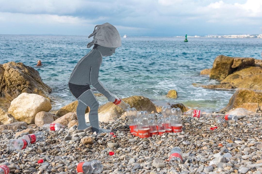 Bord de mer. Silhouette d'enfant en carton, qui ramasse des bouteilles de coca cola, plantées dans le sable et signées TOO LATE.