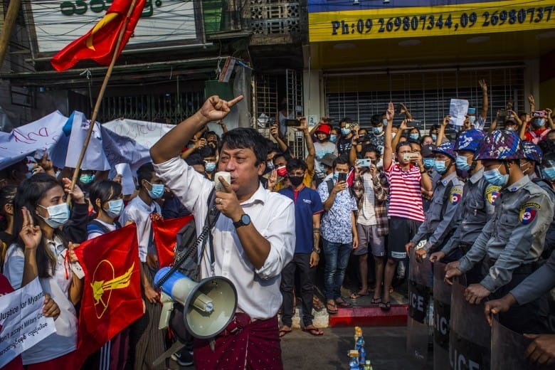 Photographie prise pendant une manifestation birmane. Un homme tient un mégaphone et s'adresse à la foule. Face à lui, un mur de policiers.
