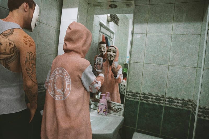 dans une salle de bain devant un miroir, deux individus avec le masque d'anonymous se montrent leur téléphone