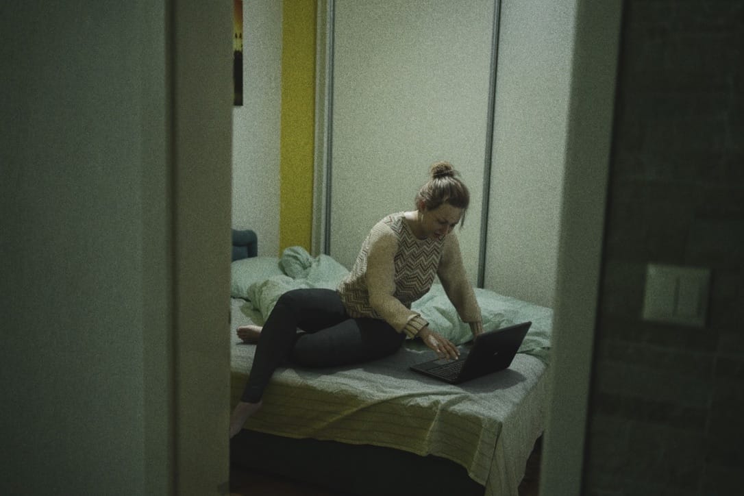 dans une chambre, sur son lit, se tient une femme assise qui consulte son ordinateur portable
