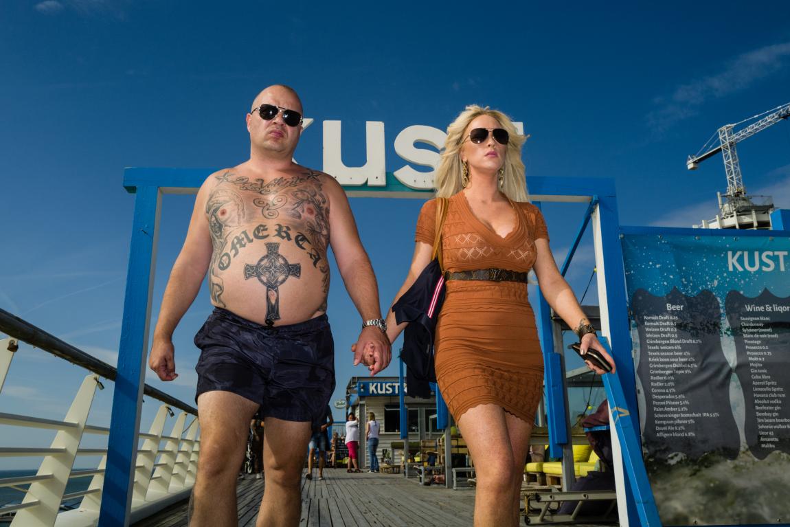 en contre plongée, un couple avec des lunettes de soleil se tient la main. Ils sont au bord de la plage, lui en maillot et elle en robe