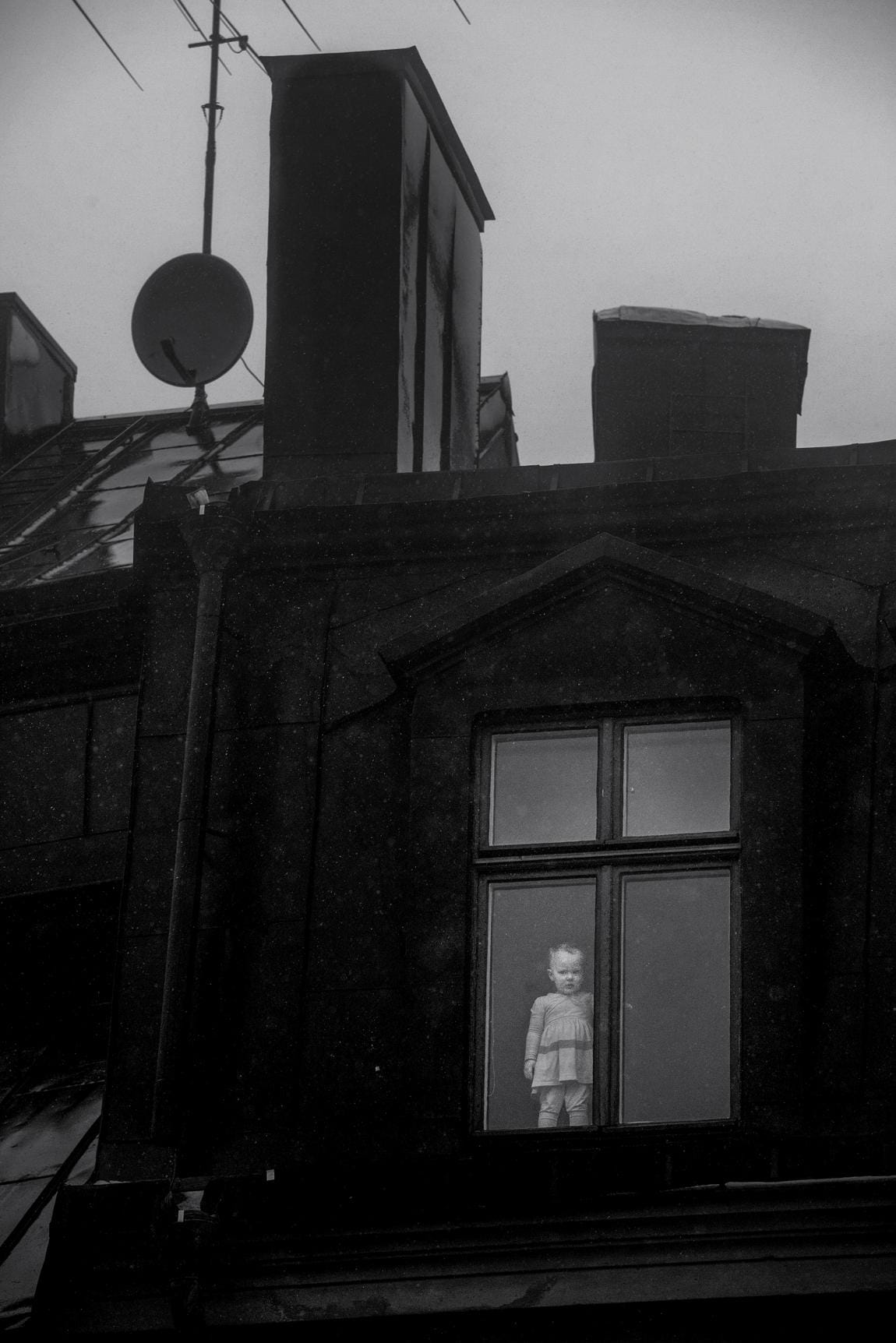 dans l'encadrure d'une fenêtre dans un cliché en noir et blanc se tient un jeune enfant, debout devant la fenêtre