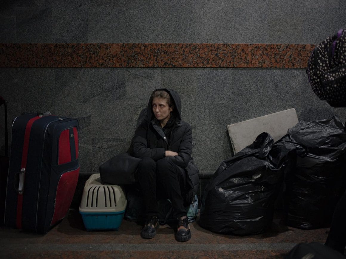 À Lviv, une femme enveloppée dans son manteau, capuche sur la tête, est assise sur un sac. Elle est entourée de bagages et d'une caisse. 