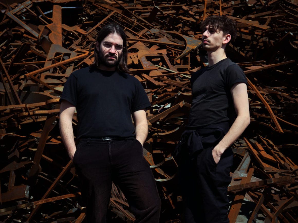 Antoine Talon et Gabriel Renault forment le groupe ATOEM. Ici, ils posent en tenue noire devant des pièces métalliques