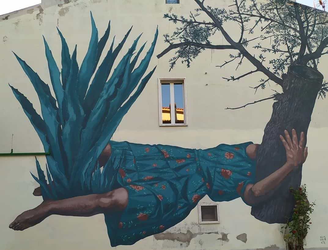 Peinture murale de Nicola Alessandrini d'une femme en robe bleue et aux motifs florales rouge faisant pont entre une plante et un arbre. 