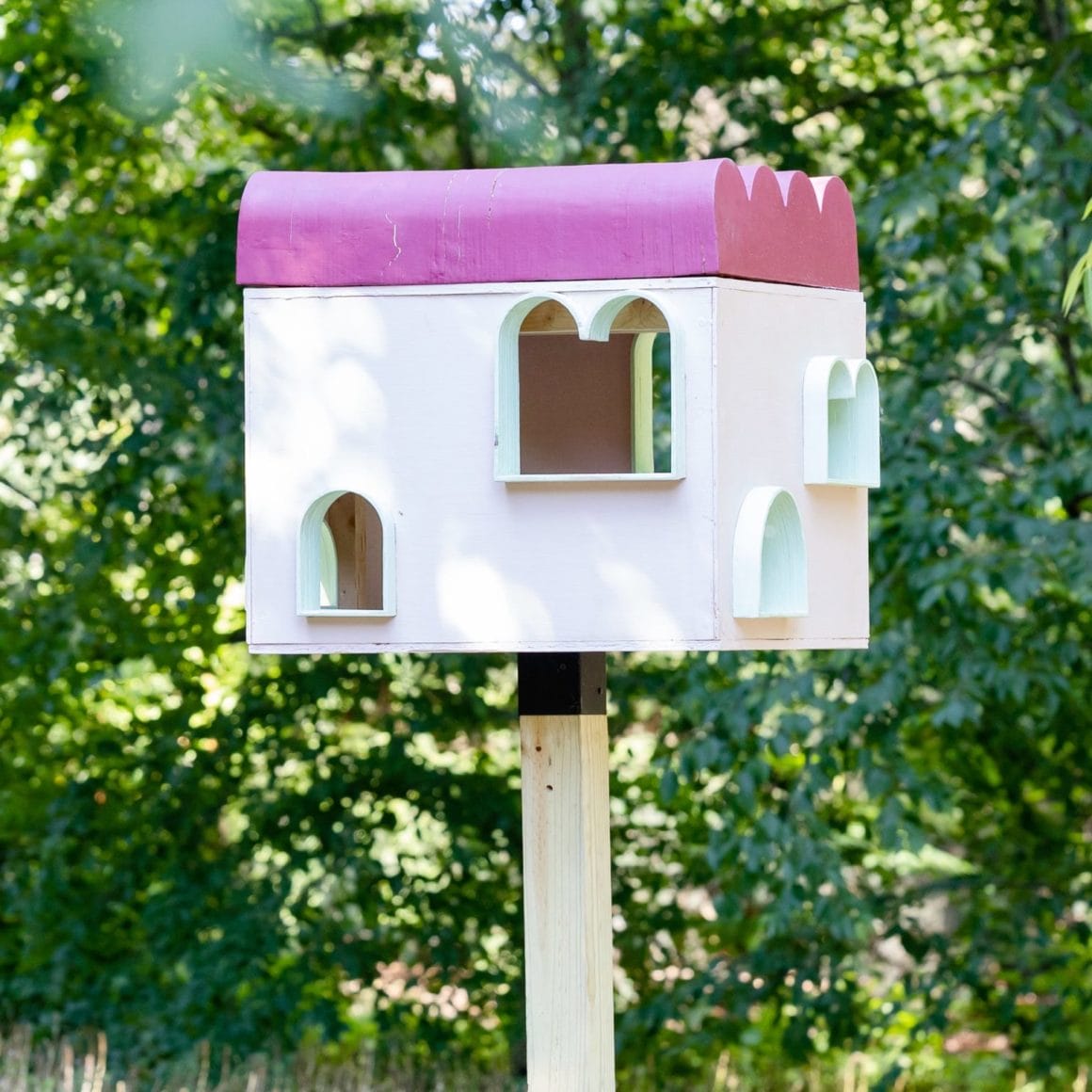 cet abri est fait de bois, avec le toit peint en rose et des "fenêtres" arrondies pour que ce soit plus facile pour les oiseaux de rentrer.