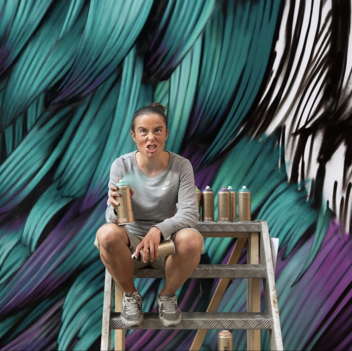 Adèle est assise sur un échafaudage en face de sa fresque murale représentant des plumes bleues, violettes et vertes