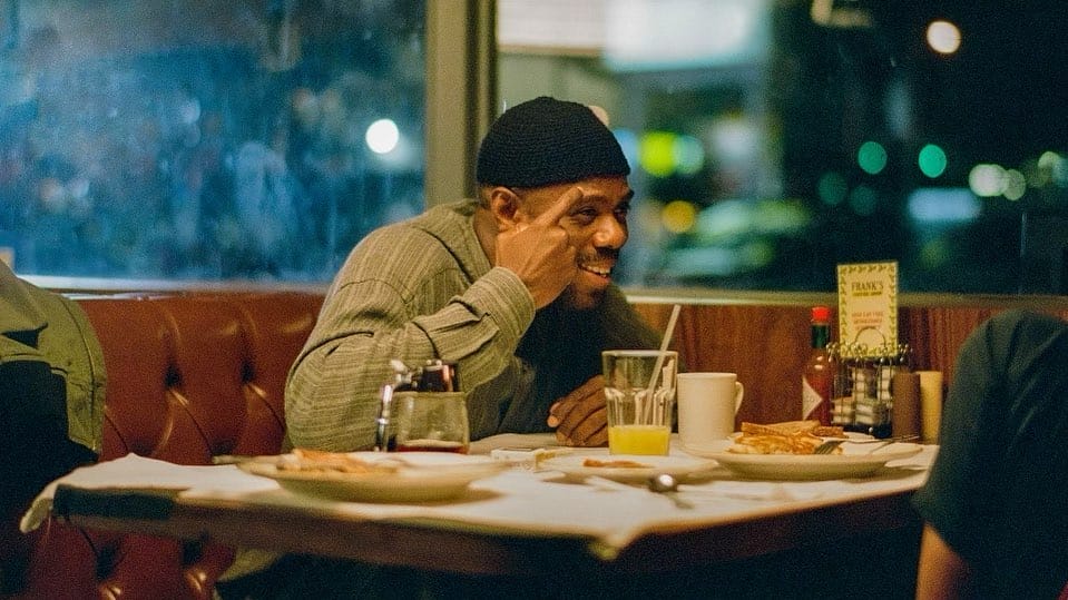 Colman Domingo est assis sur une table avec un déjeuner en face de lui. Il parle à Zendaya en souriant avec un doigt sur la tempe.