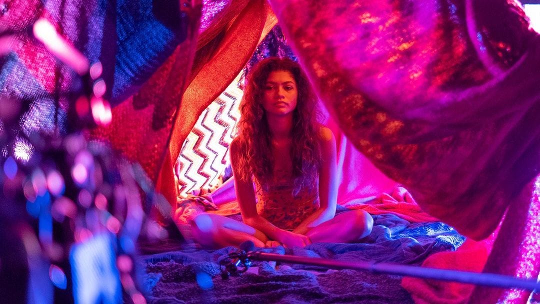 Zendaya est assise sur un lit avec des couvertures suspendues au dessus d'elle. La photo est rose bleue et violette