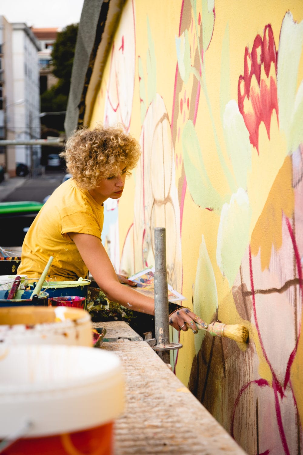 Une femme blonde aux cheveux bouclés courts peint une fresque  sur un échafaudage