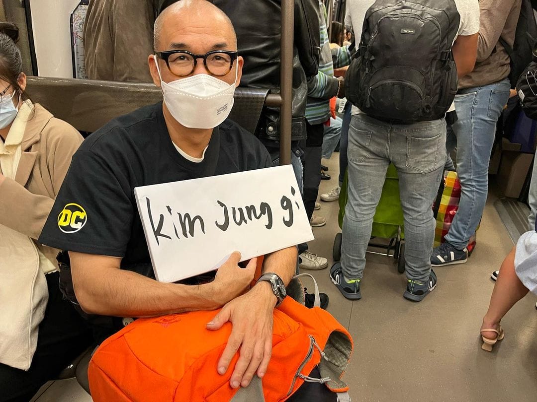 Photo de Kim Jung Gi dans le métro  de Séoul en direction de la France. Il tient une affiche avec inscrit dessus son nom.