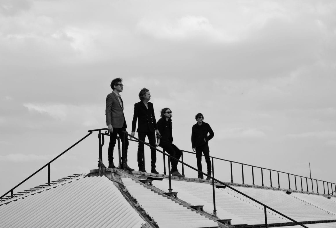 Photo en noir et blanc de quatre homme en costume noir installés en haut de marches