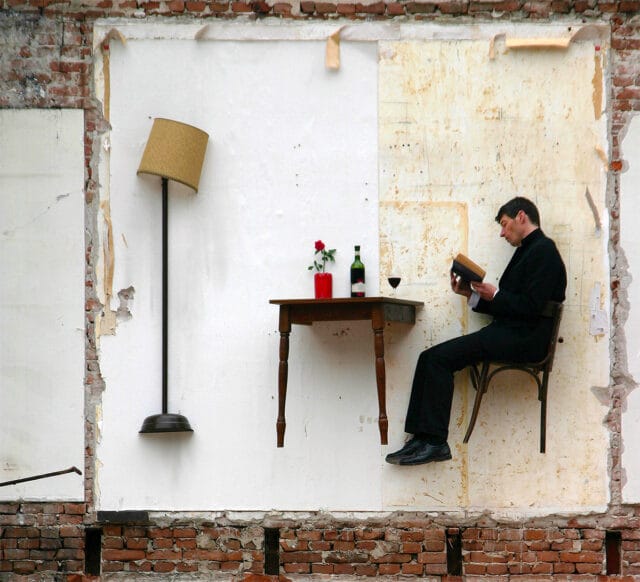 Sur une façade délabrée est accroché une table et une chaise coupée, sur lesquelles un homme lit un livre.