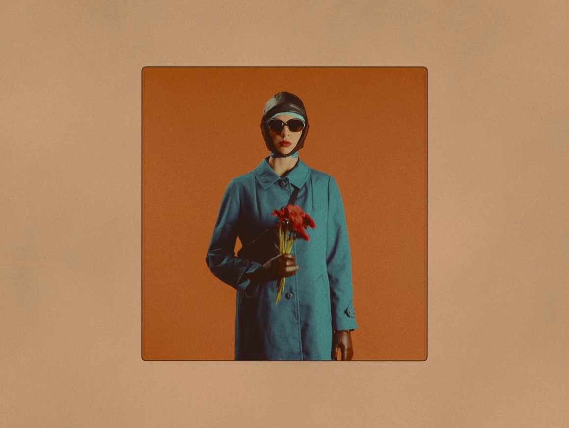 Marion Tiratay dans un manteau bleu sur fond orange, dans un cadre, avec bouquet de fleurs, lunettes de soleil et casque